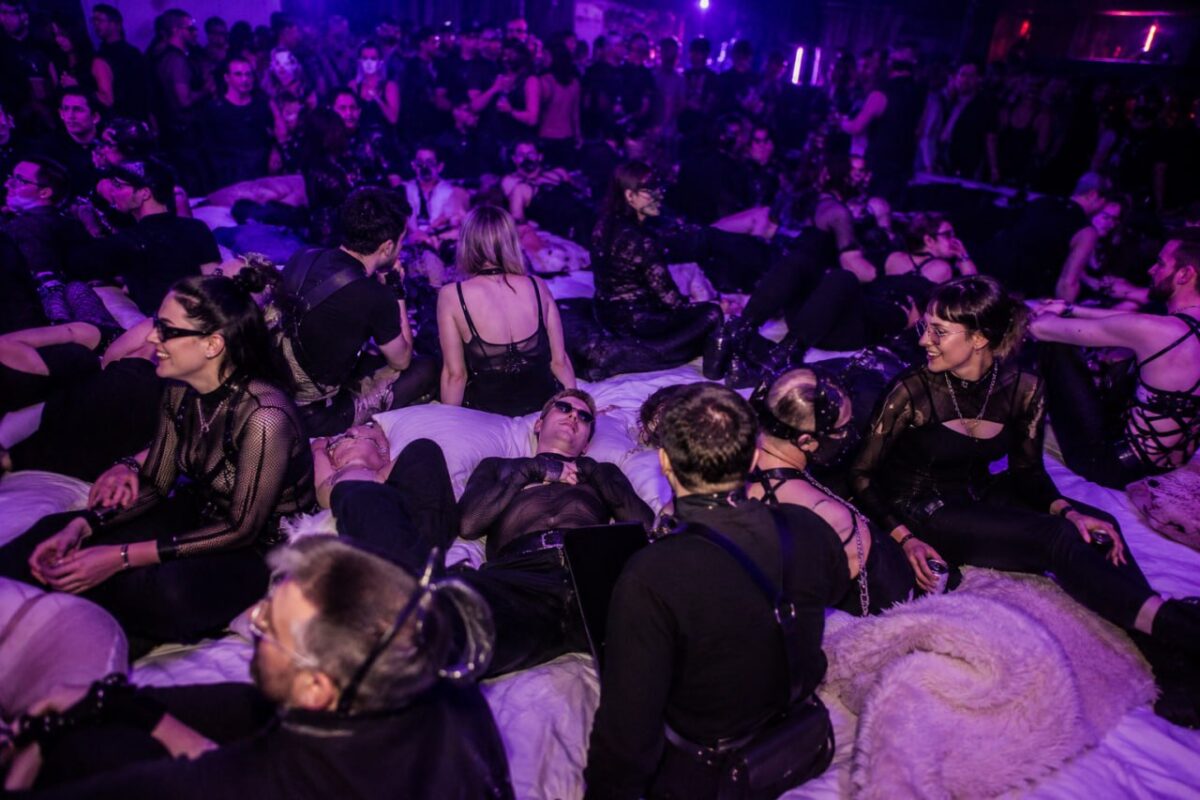 Young couples at a purple-lit lifestyle party, gathered on lounge beds in black fetish clothing