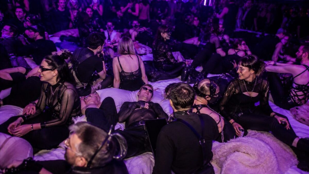 Young couples at a purple-lit lifestyle party, gathered on lounge beds in black fetish clothing